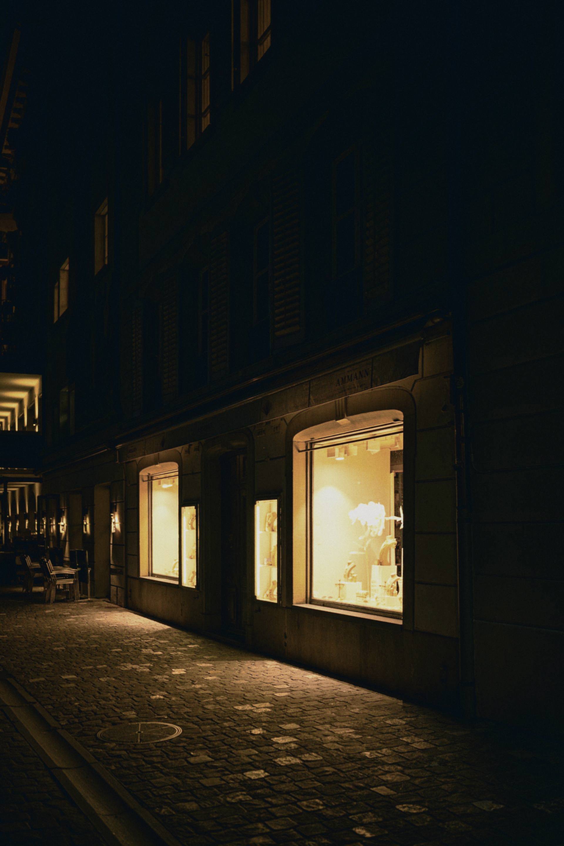 A shop window still lit after closing on an empty street, no sign legible.